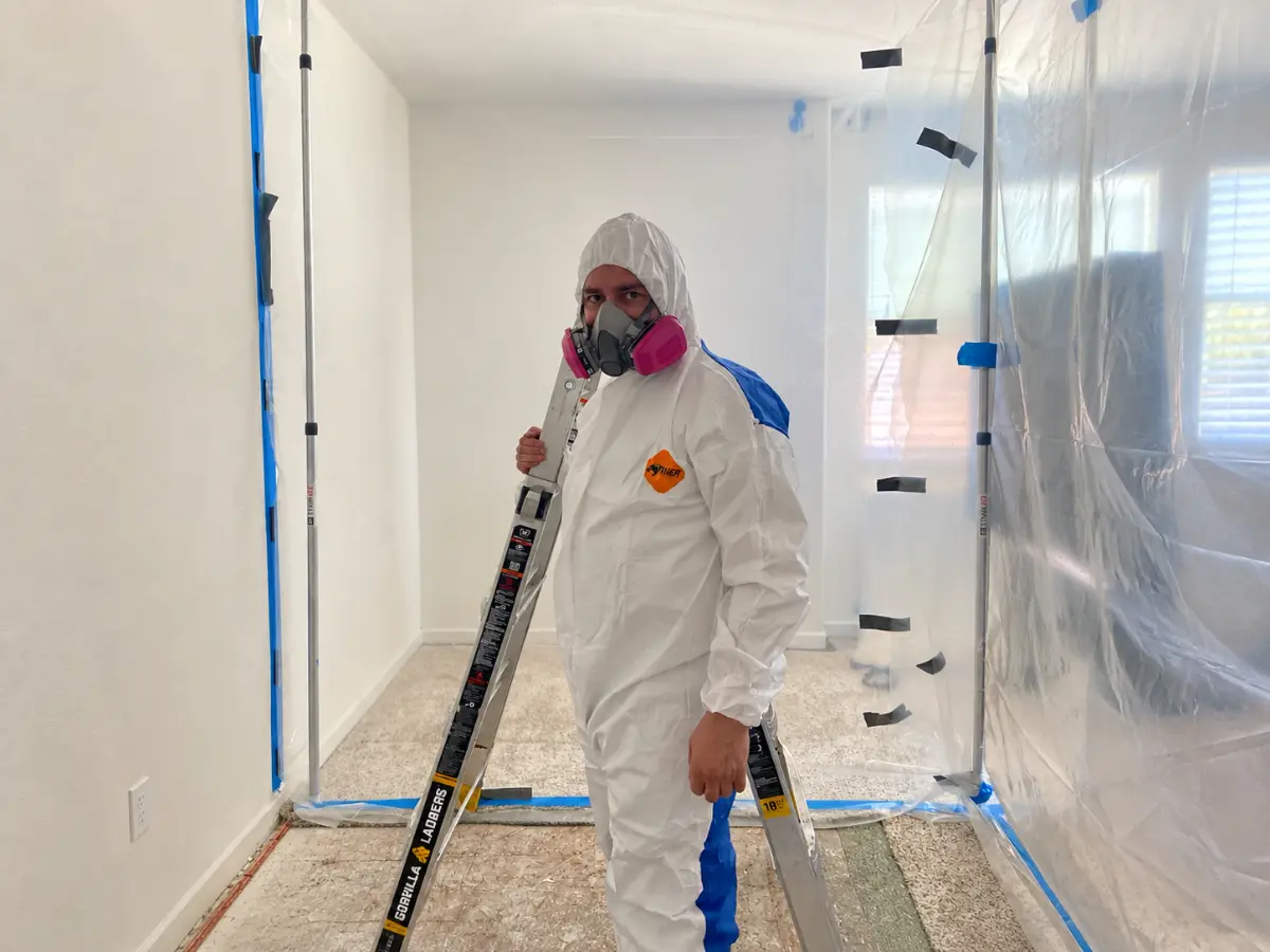Restoration technician wearing a protective suit and respirator inside a plastic containment area with a ladder.